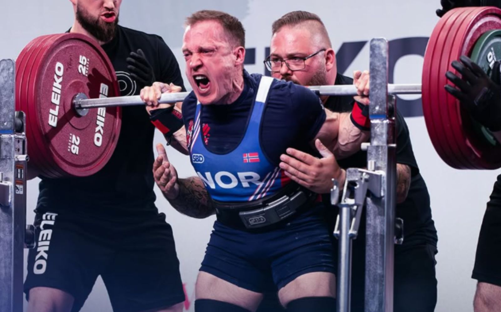 Atleta de powerlifting ergue barra com discos coloridos sob iluminação de arena, enquanto tela exibe logo da IPF