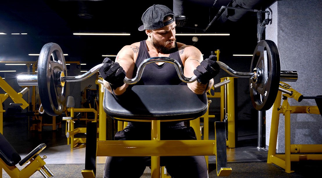 Um homem de braços musculosos fazendo preacher curl em uma máquina de ginásio, com o assento ajustado para posição correta