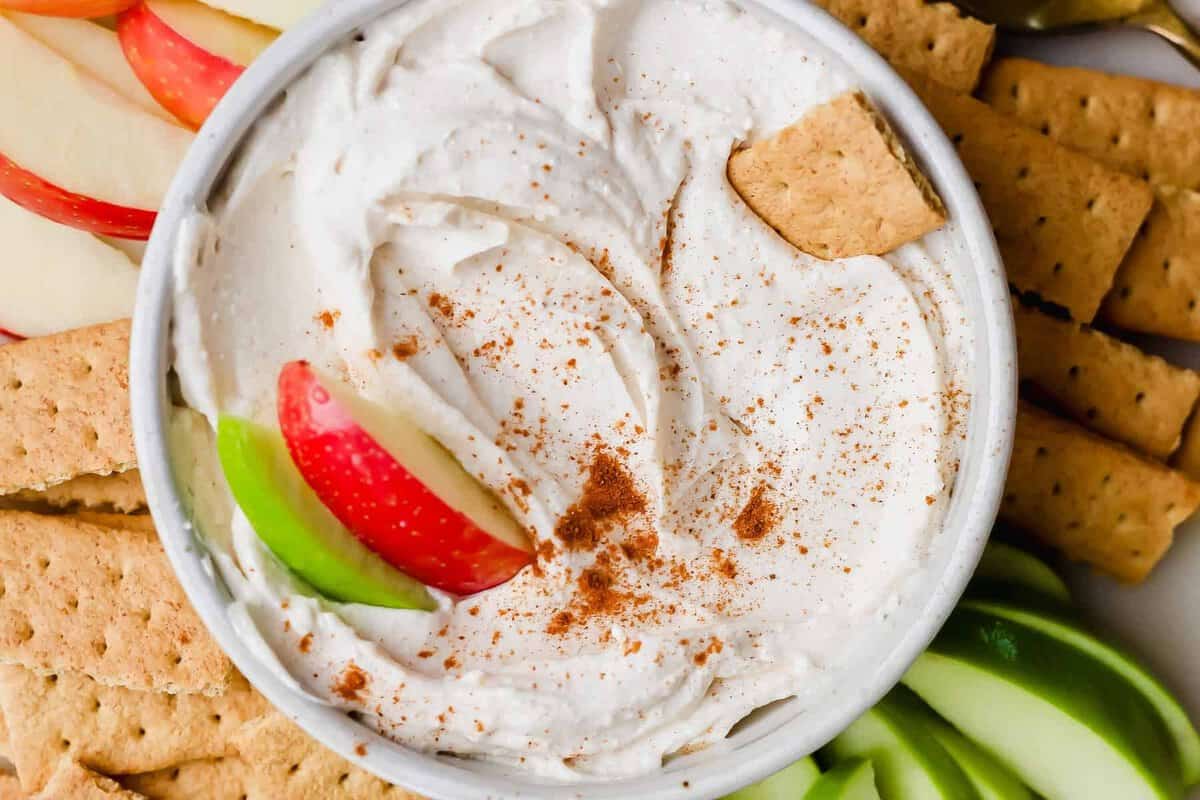 Uma maçã cortada em fatias, um prato de cream cheese e cottage cheese ao lado, com um garfo e um pão de trigo
