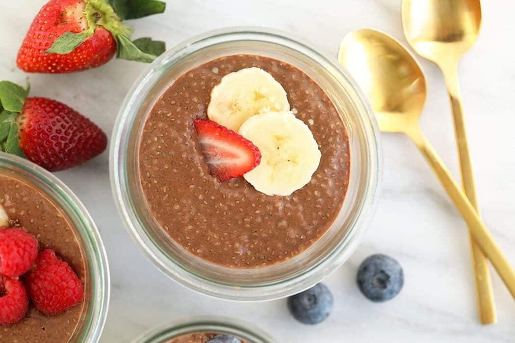 Pote de vidro com pudim de chia, frutas frescas e sementes sobre uma mesa de madeira ao lado de uma colher