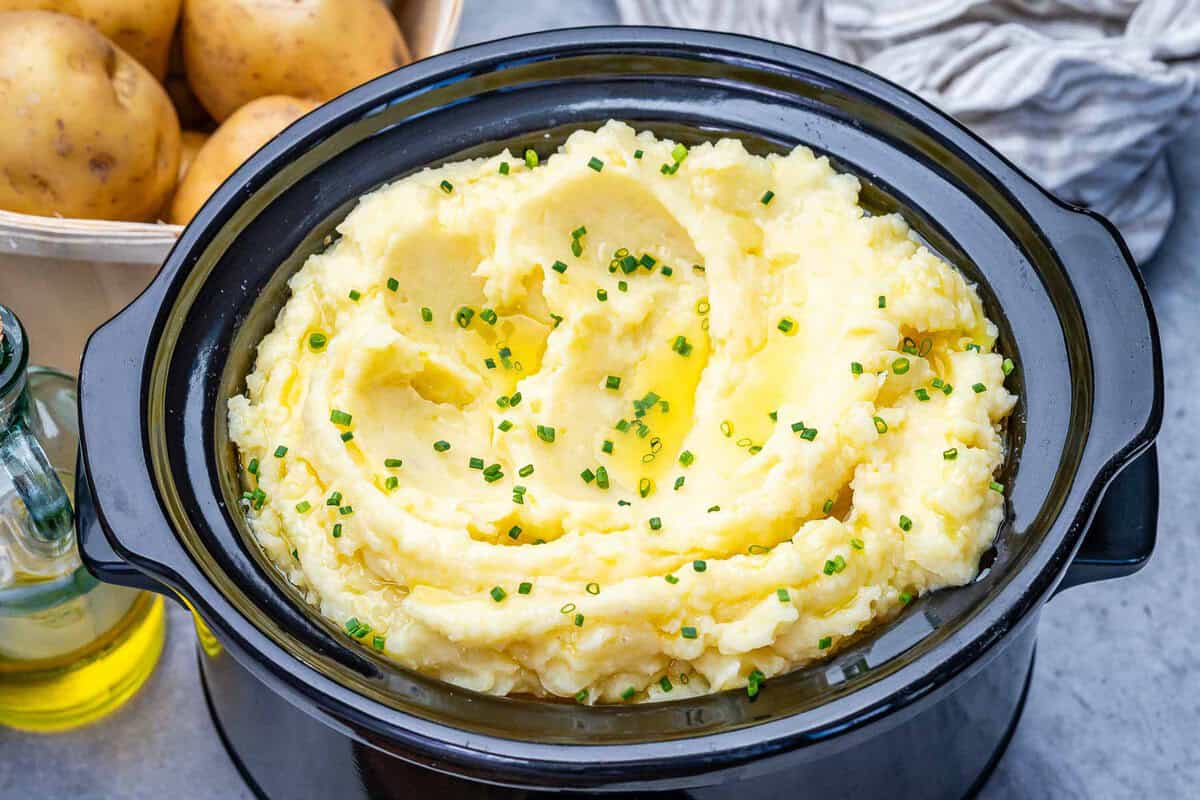 Batatas Yukon Gold, leite, parmesão e azeite na slow cooker, ao lado de colher de madeira e folhas de salsa