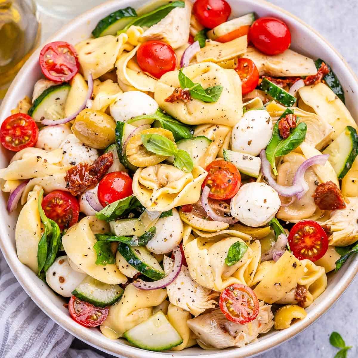 Bowl de cerâmica com salada de tortellini, tomates cereja, espinafre fresco e azeite sobre mesa de madeira clara