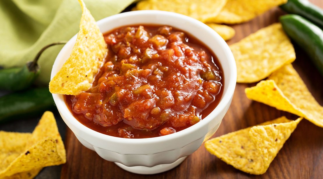 Frasco de salsa industrial ao lado de tomates, cebola roxa, pimentão verde e limão, com rótulo destacando baixo teor de sódio