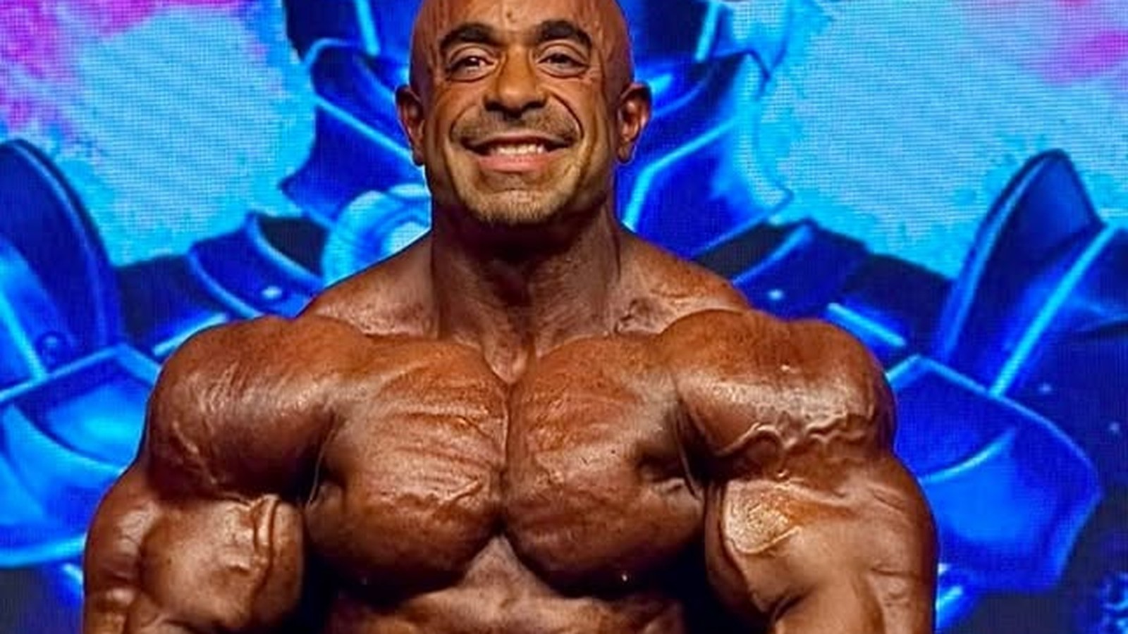 Sasan Heirati exibindo seu físico musculoso e definido em pose clássica de fisiculturismo sob luzes de palco