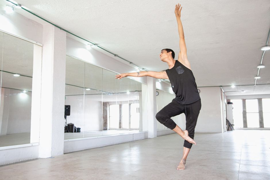 Mulher em roupa esportiva, segurando halteres leves, executando um plié na barra de ballet, com espelho e luzes de estúdio ao fundo