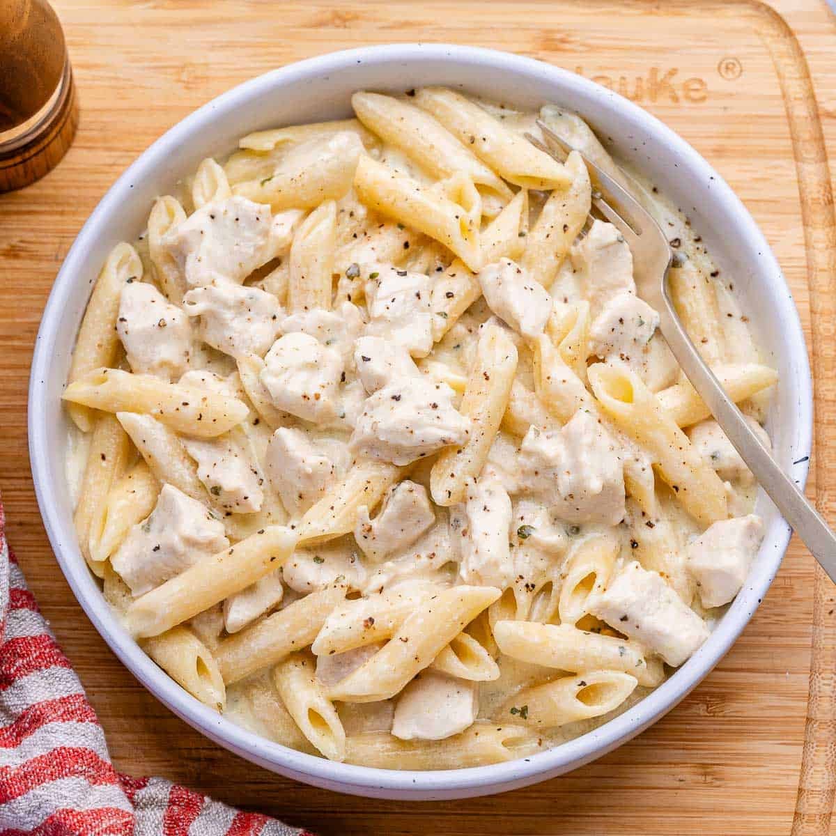 Um prato de pasta com frango cozido em molho alfredo em uma panela lenta, com um copo de proteína em pó ao lado