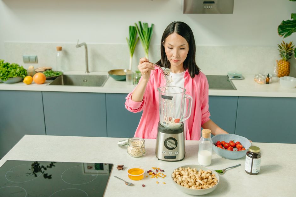 Copo transparente com smoothie verde, pedaços de maçã e abobrinha ao lado, colher e liquidificador ao fundo