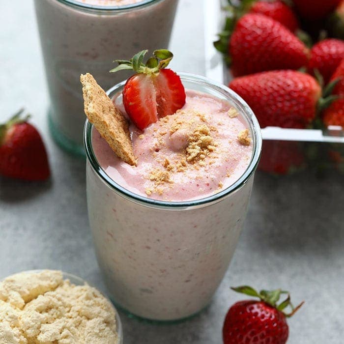 Um copo de smoothie vermelho com morangos e batata-doce, acompanhado de um copo de proteína em pó e um garfo