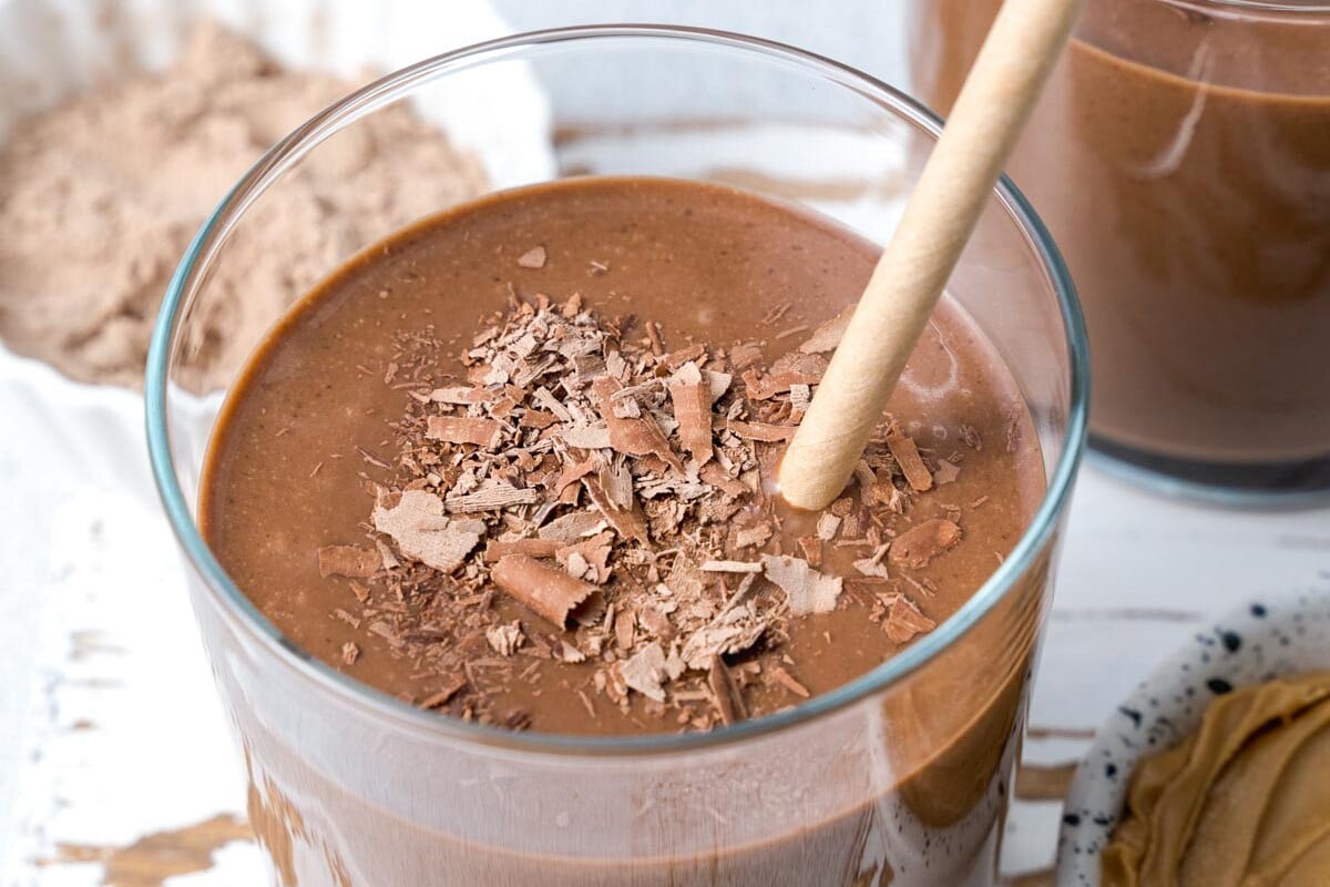 Pote de whey protein aberto sobre bancada de cozinha ao lado de uma coqueteleira e frutas frescas para o pós-treino