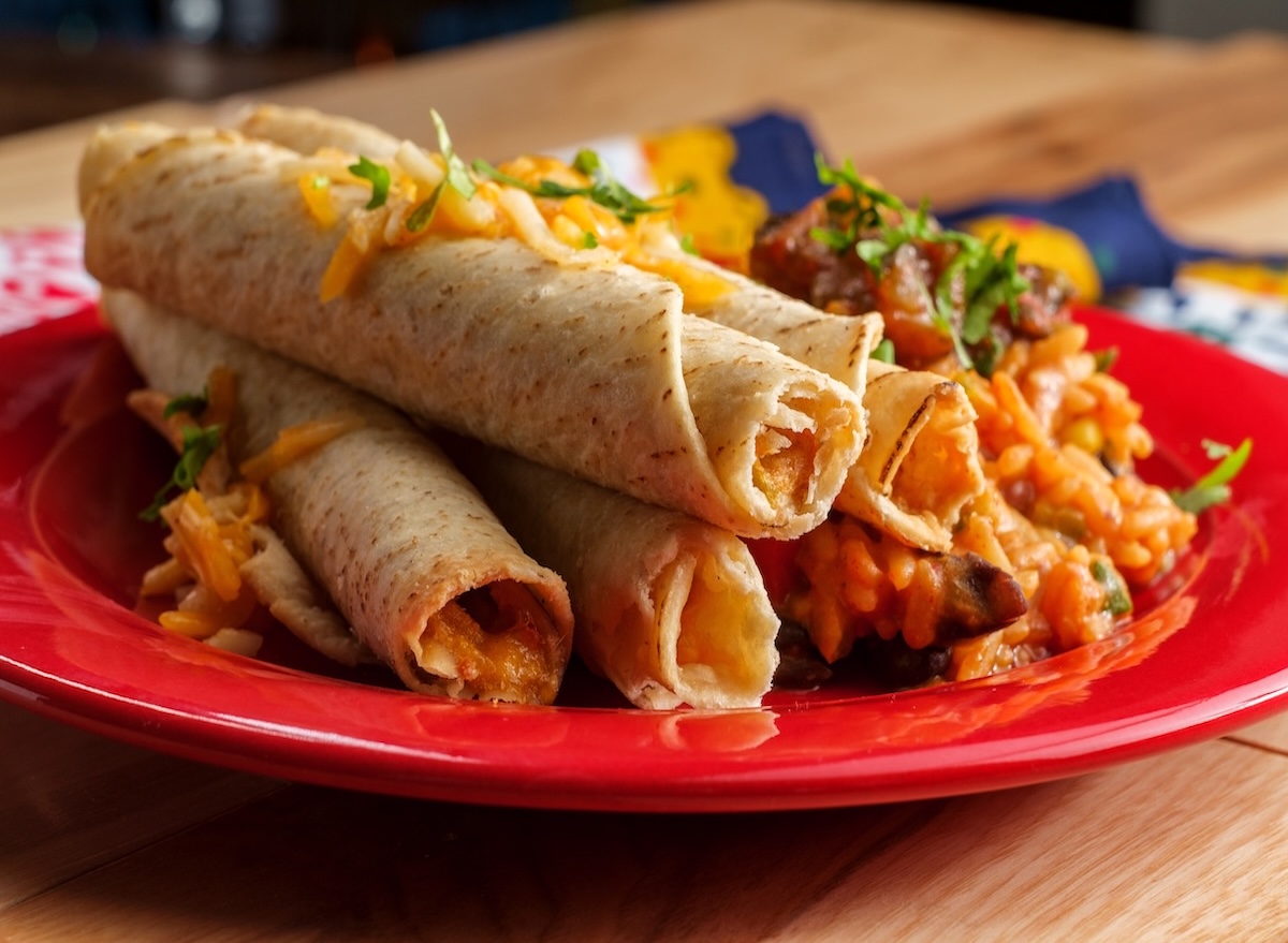 Taquitos dourados e crocantes dispostos em uma tábua de madeira, acompanhados de molho guacamole e limão fresco