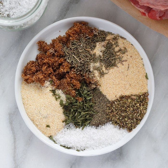 Pote de tempero caseiro ao lado de carne assada suculenta, legumes grelhados e uma tábua de corte com ervas frescas