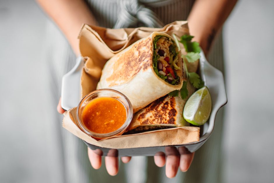 Burrito enrolado com tofu dourado, feijão preto, espinafre verde e molho, ao lado de uma tábua com limão e pimenta