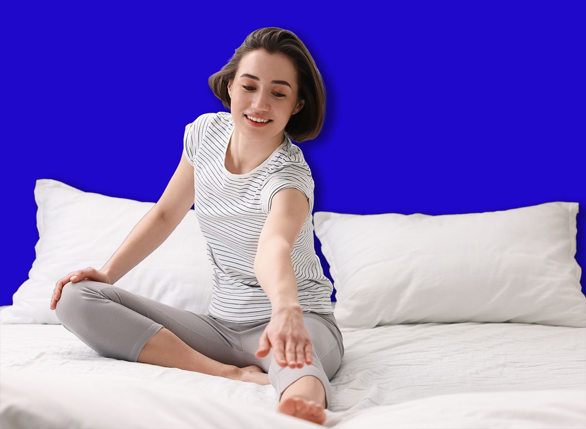 Uma mulher sentada na cama, com as costas retas, realizando um dead bug, com um livro ao lado e uma garrafa de água na mesa de cabeceira
