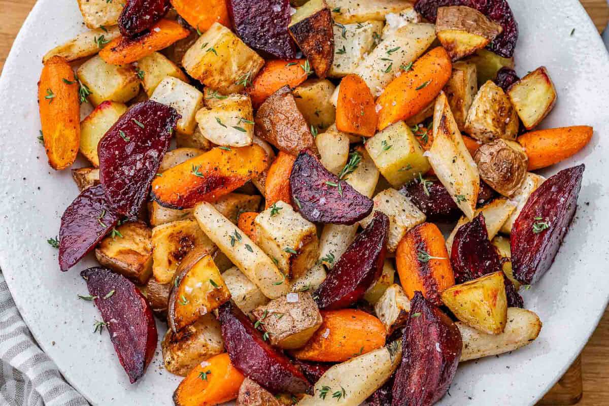 Batatas, beterrabas, cenouras e nabos assados, dourados, dispostos em uma assadeira com alecrim e azeite