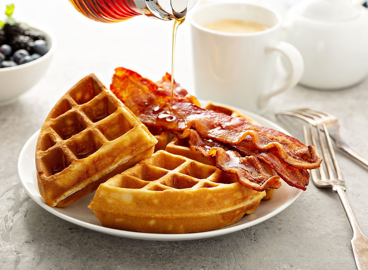 Waffles dourados empilhados ao lado de tiras de bacon crocante, com mel escorrendo e uma xícara de café ao fundo