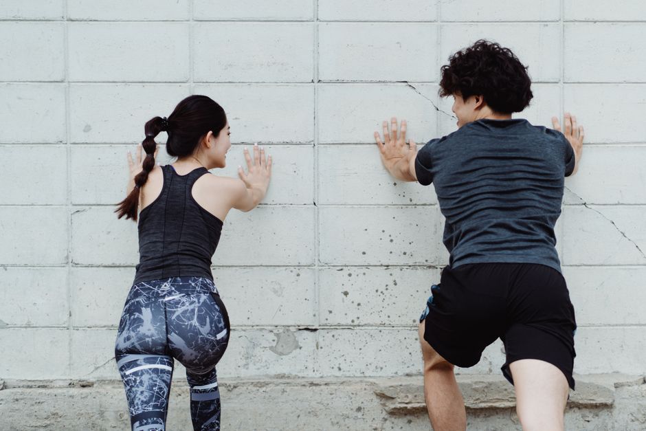Uma pessoa sentada com as costas contra uma parede, com as pernas estendidas e os pés afastados, em uma pose de força de pernas